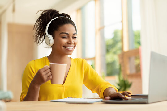 Happy african american lady in headphones working on laptop, holding cup and drinking coffee, sitting at desk at home