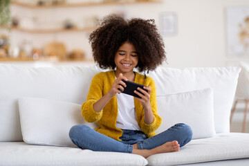 Happy african american little girl sitting on couch with smartphone