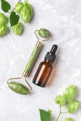 Unbranded serum bottle, face roller with hop on marble background. Jade face roller for facial massage therapy. Anti age, lifting and toning treatment