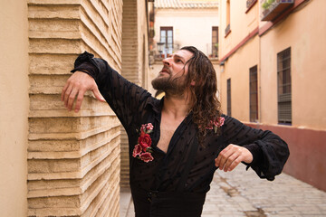Long haired man dancing flamenco with black shirt and red roses. He makes dancing postures with his hands in a typical narrow street of Seville. Flamenco dance concept cultural heritage of humanity. - obrazy, fototapety, plakaty