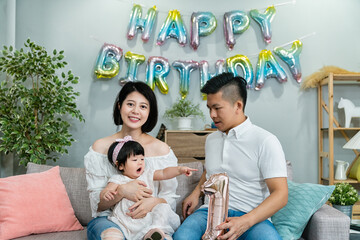 Asian baby girl showing interest in the number standee her dad is holding by finger pointing at it while her mom is smiling at camera on birthday celebration
