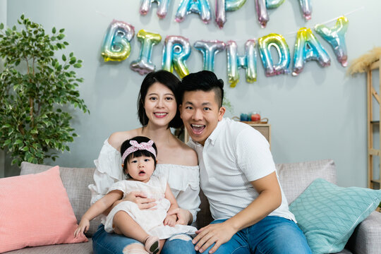 Sweet Asian Nuclear Family With A Lovely Baby Smiling At The Camera While Celebrating Birthday In A Modern Bright Living Room With Festive Decoration At Home