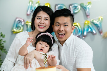 adorable asian baby girl is looking at the cake with curiosity while her parents is smiling at the camera and celebrating her first birthday indoors
