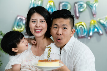 cute asian baby girl having fun looking at her dad who’s blowing candles on cake while they are taking family photo on the occasion of her first birthday at home