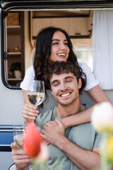Cheerful woman hugging brunette boyfriend with wine near camper van