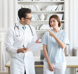 Front view on male doctor and female patient standing in front of the desktop in the office and talking about illness