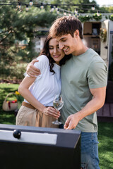 Smiling man hugging girlfriend with wine near blurred grill outdoors