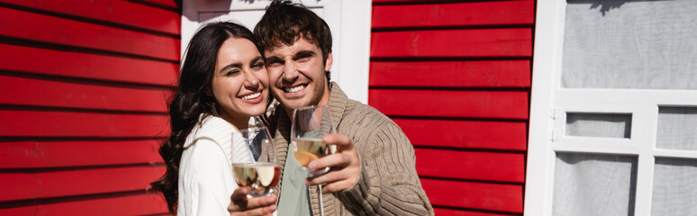 Cheerful young couple holding glasses of wine and looking at camera near house, banner