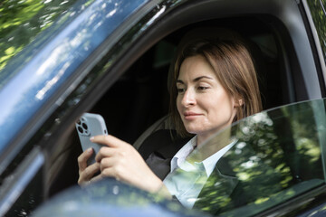 Young and attractive business woman in black suit sitting in the car and holding smartphone. Serious woman driving and looking at phone