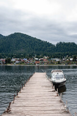 A water boat is moored at an old pier made of wooden boards on a mountain lake. Mountains and village in the background