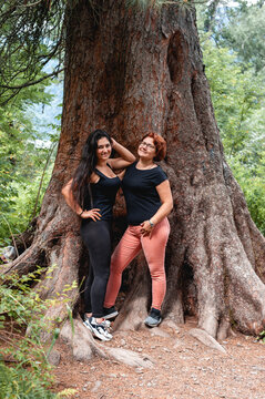 two adult cute girls of different races, Caucasian and Middle Eastern, standing near a thick tree trunk. Women with different hairstyles and hair colors. In black t-shirts