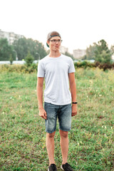 Fototapeta premium A cheerful Caucasian guy with glasses and a cap on his head is dressed in a white T-shirt and jeans made a grimace on his face, stretching his lips. Summer photo at sunset