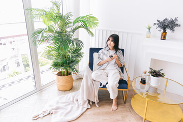Young pretty woman smile and sits in the chair of the modern living area with a green plant, Relaxing drinking a cup of hot coffee, Healing reflection in an indoor environment at a cozy home.