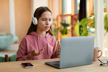 Online communication. Teenage girl talking at laptop webcamera, wearing wireless headphones