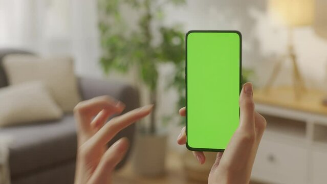 Handheld Camera: Back View of Young Woman at Home Living Room Holding Chroma Key Green Screen Smartphone Watching Content With Touching or Swiping Left Three Times..