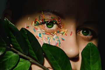 An open, green woman's eye with a sweet, multicolored sprinkle on the eyelid and a plant with...
