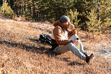 Young man in autumn clothes drinking tea from a thermos while sitting at the edge of the forest in evening sun resting in hike and enjoying nature