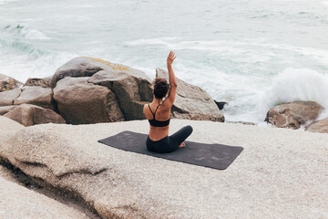Rear view of fit female raising hand while changing yoga pose. Woman sitting on mat by shore and...