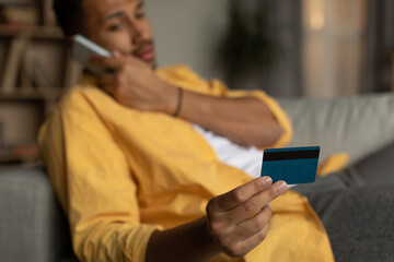 Young black man holding credit card, calling to bank client service, having problem during online shopping at home
