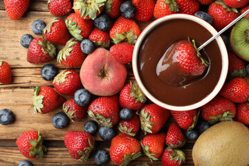 Fondue fork with strawberry in bowl of melted chocolate surrounded by other fruits on wooden table, flat lay