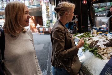 Portrait of two mature female friends standing in front of a fish shop looking apparently at the...