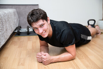 A young teen guy, does sports at home, stands in static plank position, strengthens his back, legs, and abdominal muscles. The guy is smiling and standing in the plank, after aerobic exercise