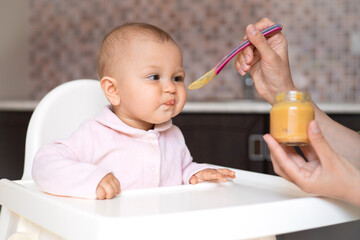 Baby food. A baby in a high chair eats vegetable puree from a spoon. Mom feeds the baby from a jar. Kitchen. Lifestyle.