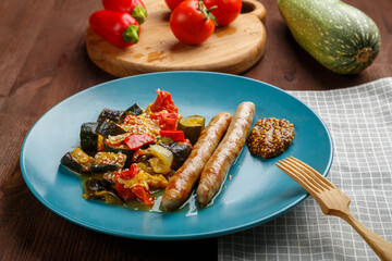 A plate of vegetables with grilled sausages on the table next to zucchini and tomatoes on wooden boards and a wooden fork.