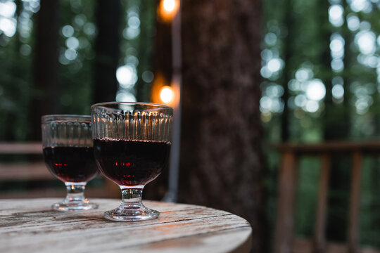 Two glasses of red wine standing on the wooden table with blurred trees and lights on the background.