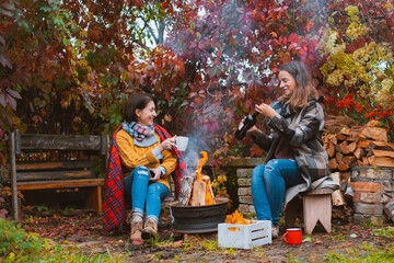 two happy joyful best friends sitting outside in autumn garden by campfire with bottle of red wine
