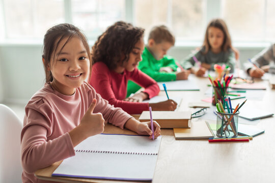 Asian Schoolgirl Gesturing Thumbs Up Smiling Taking Notes In Classroom