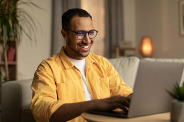 Beautiful young black man working on laptop pc, enjoying remote job or education in living room