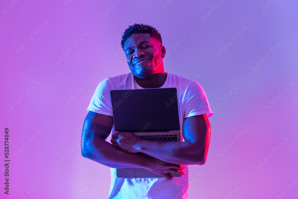 Poster cheerful young black man hugging laptop with empty screen in neon light, mockup for website