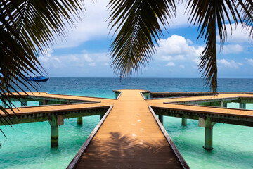 Wooden walkways on the pier of a resort island with sand marks in the Maldives against the background of hanging palm leaves, a sky with clouds and clear turquoise water of the ocean and a boat