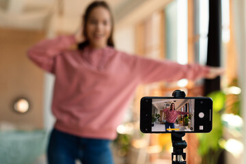 Modern teenage female influencer recording video on smartphone, dancing on camera at home,...
