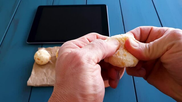 Man taking Brazilian cheese rolls and splitting them in half next to a tablet and coffee cup. Blue wood background.