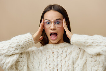 a happy, mischievous woman in eyeglasses stands with her mouth wide open with happiness and holding her glasses with her hands while standing on a beige background. 