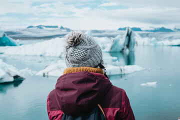 Traveler woman in cold weather clothes in front of the glaciers of Jökulsárlón in Iceland during a moody day filled with water. Live your dream, love in Iceland, road trip style.Copy space - obrazy, fototapety, plakaty