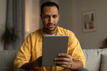 Focused young black man with tablet sitting on sofa, working or studying online, having remote business meeting at home