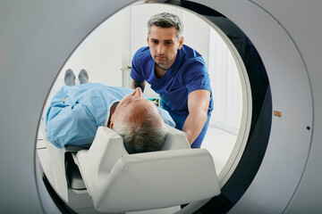Senior man going into CT scanner. CT scan technologist overlooking patient in Computed Tomography scanner during preparation for procedure - obrazy, fototapety, plakaty
