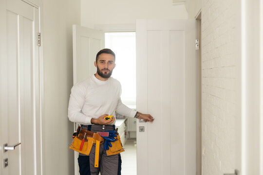 Installation Of A Lock On The Front Wooden Entrance Door. Portrait Of Young Locksmith Workman In Blue Uniform Installing Door Knob. Professional Repair Service. Maintenance Concept