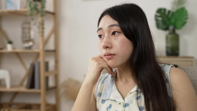 closeup view of a sad Asian female propping face and looking into distance with a sigh feeling loneliness while staying at home alone during pandemic outbreak.