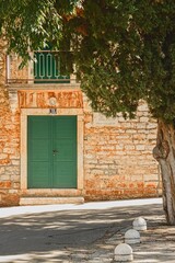 Photo of empty small street with romantic house with stone facade, wooden green door and ornamental balcony in background hidden behind huge tree. Town Supetar, Croatia, Europe