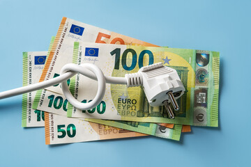 Knotted power cable with electric plug on 50 and 100 euro banknotes over blue background. Concept of rising energy costs in Europe. Electric bill increases. Power price spike.