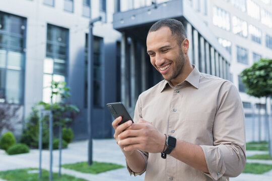 Young african american businessman using smartphone, smiling and happy typing message online, man outside office building typing message to friends.