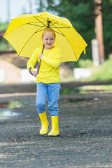 portrait of a 7-year-old girl in yellow clothes and with a bright yellow umbrella in her hands, who walks through the puddles in rubber boots in the warm season