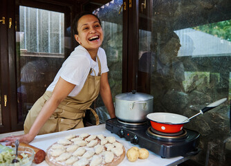 Stunning woman housewife in chef's apron, standing by table with raw dumplings sprinkled with flour...