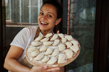 Pretty woman in beige chef's apron, housewife smiling at camera, holding a wooden board full of...