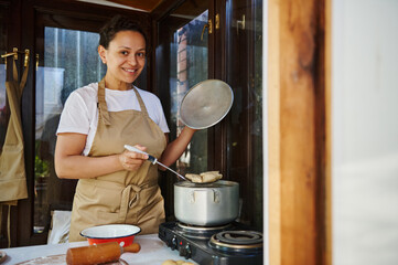 Charming woman housewife in beige chef's apron, stands by stove, stirs homemade dumplings in the...