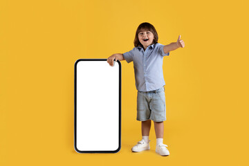 Modern technologies for kids. Happy little boy showing thumb up gesture, posing at big smartphone with blank screen